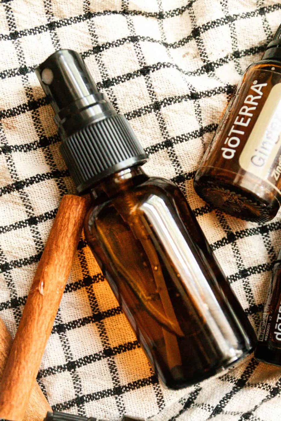 A glass spray bottle with a cinnamon stick and essential oil bottle on a checkered towel.