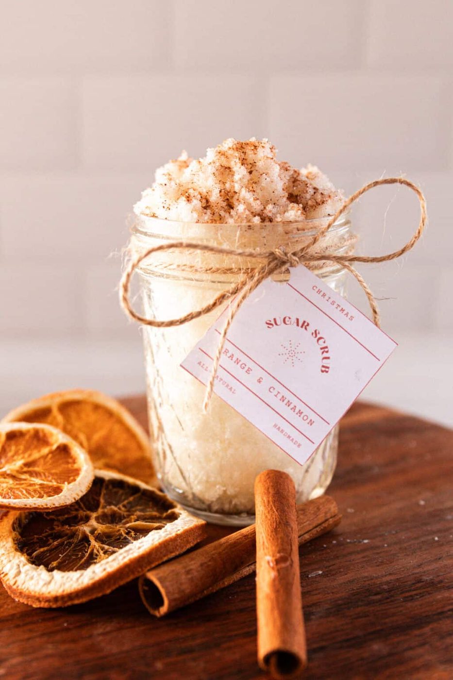 A jar of orange cinnamon sugar scrub with dried orange sliced and cinnamon sticks on a wooden board.