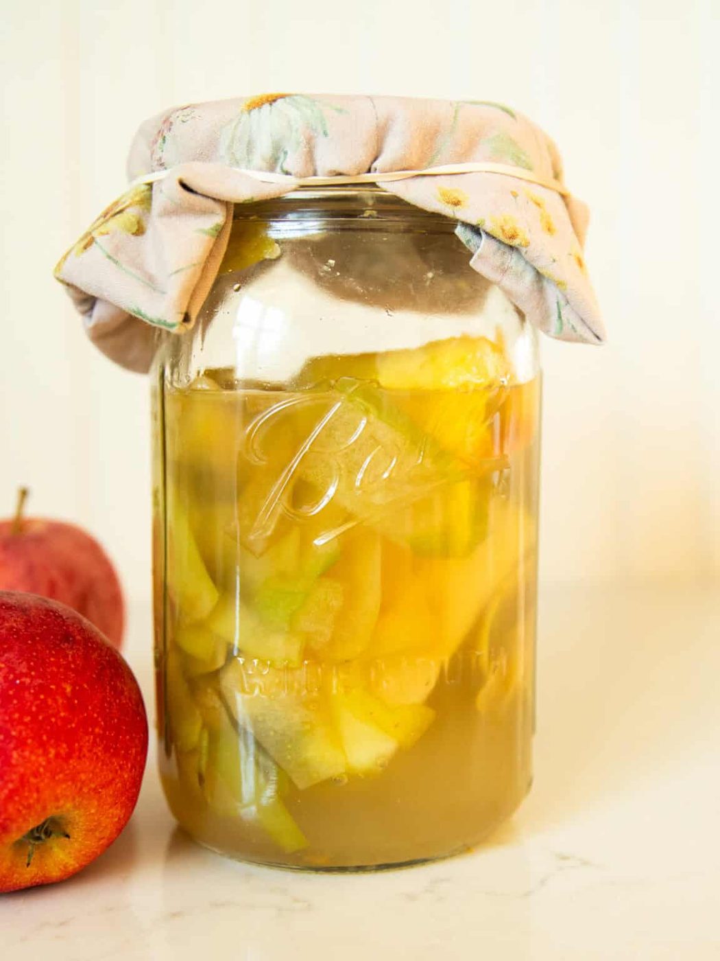 A jar of apple cider vinegar with a cheesecloth on the top and apples next to it.