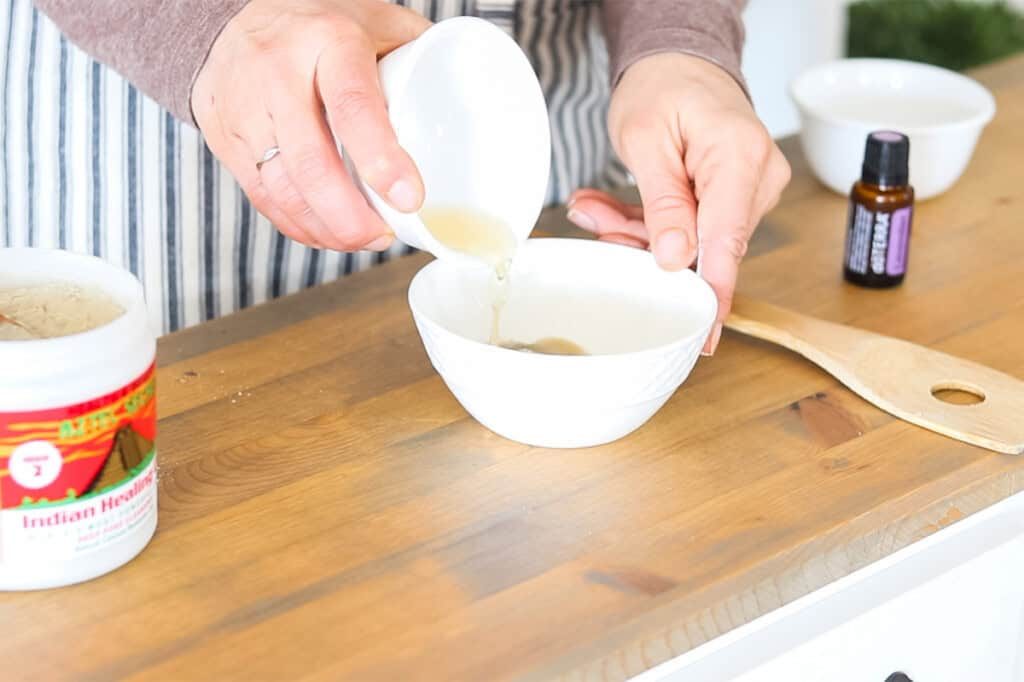 Pouring apple cider vinegar into a bowl of bentonite clay.