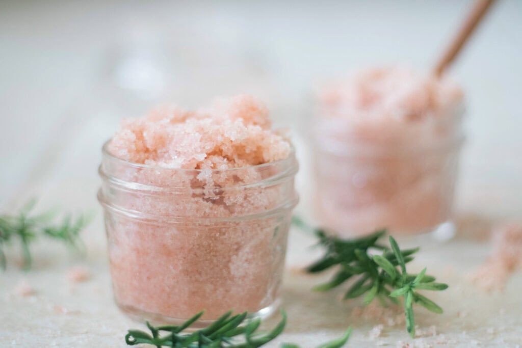 Two jars of foot scrub with fresh herbs laying around them.