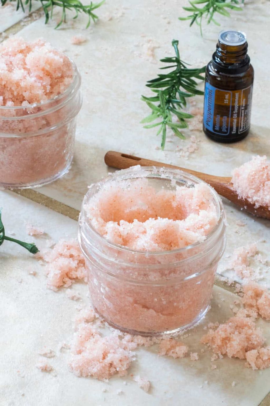 Pink foot scrub in a glass jar with another jar and a bottle of essential oil behind it.