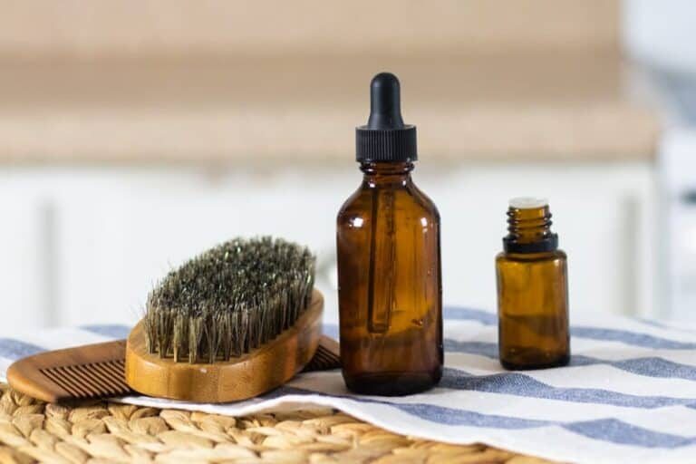 Beard shampoo on table with essential oil bottle, beard brush, and bread comb next to it.