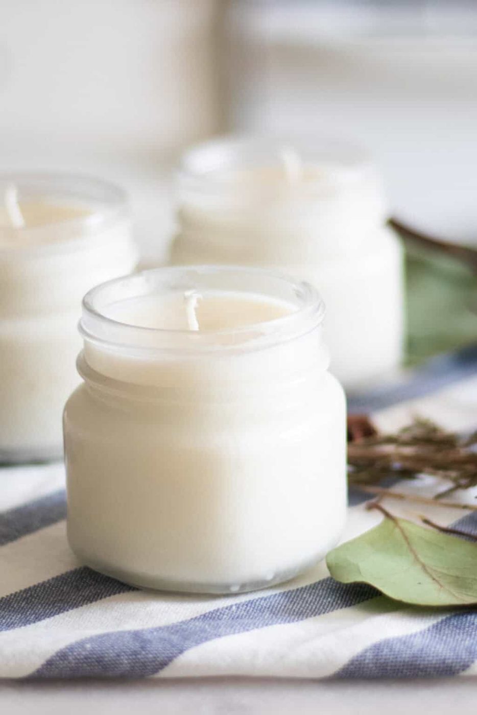 Three homemade Christmas candles with greenery on a white and blue towel.