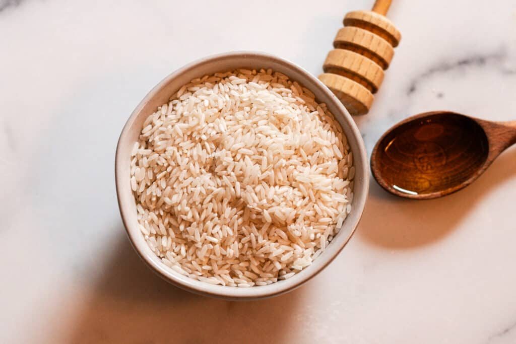 A bowl of dry rice sits on a white and gray countertop to make rice water for a DIY hair mask recipe. There is a wooden tablespoon of honey resting next to it.
