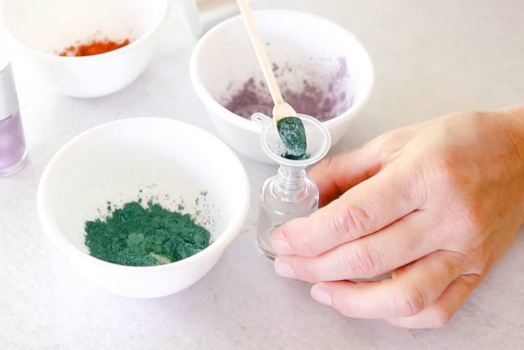Scooping teal mica powder into an empty nail polish bottle with a small funnel in the neck.