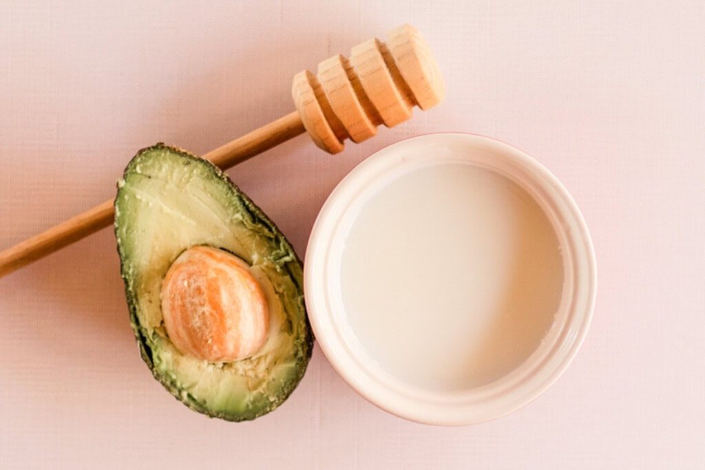 A half an avocado with a honey dipper and a bowl of milk.