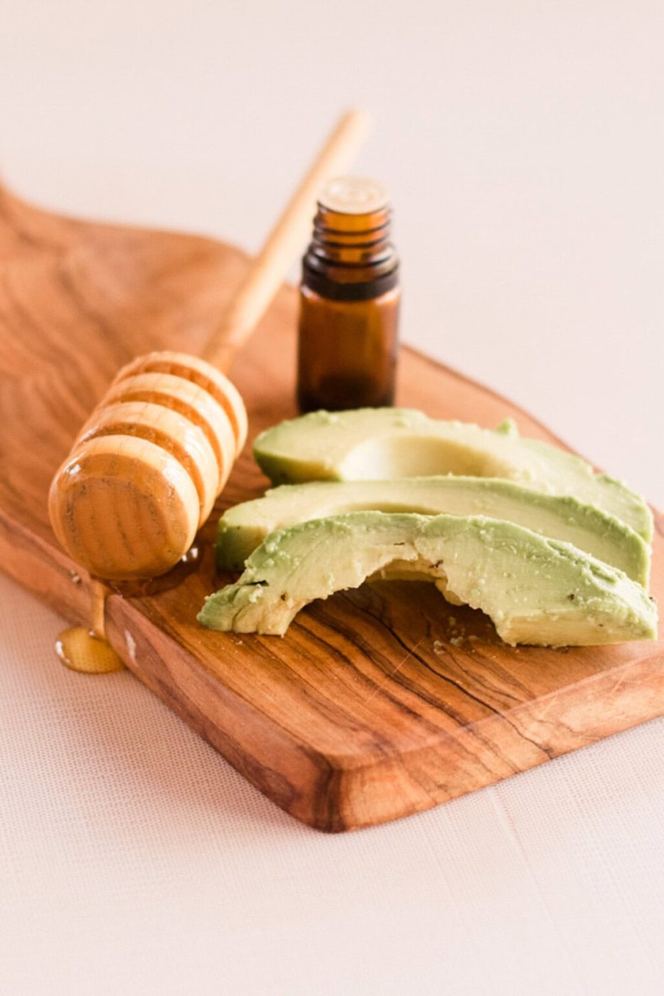Sliced avocado, a honey dipper, and a bottle of essential oil on a wooden board.