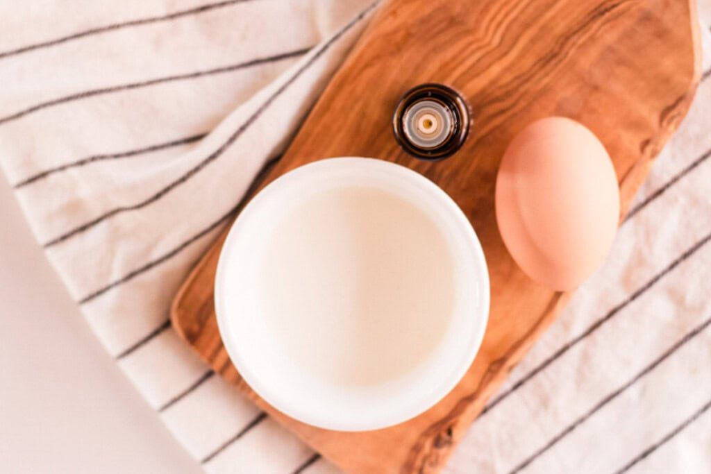 A bowl of yogurt on a wooden board with an egg and a bottle of essential oil.