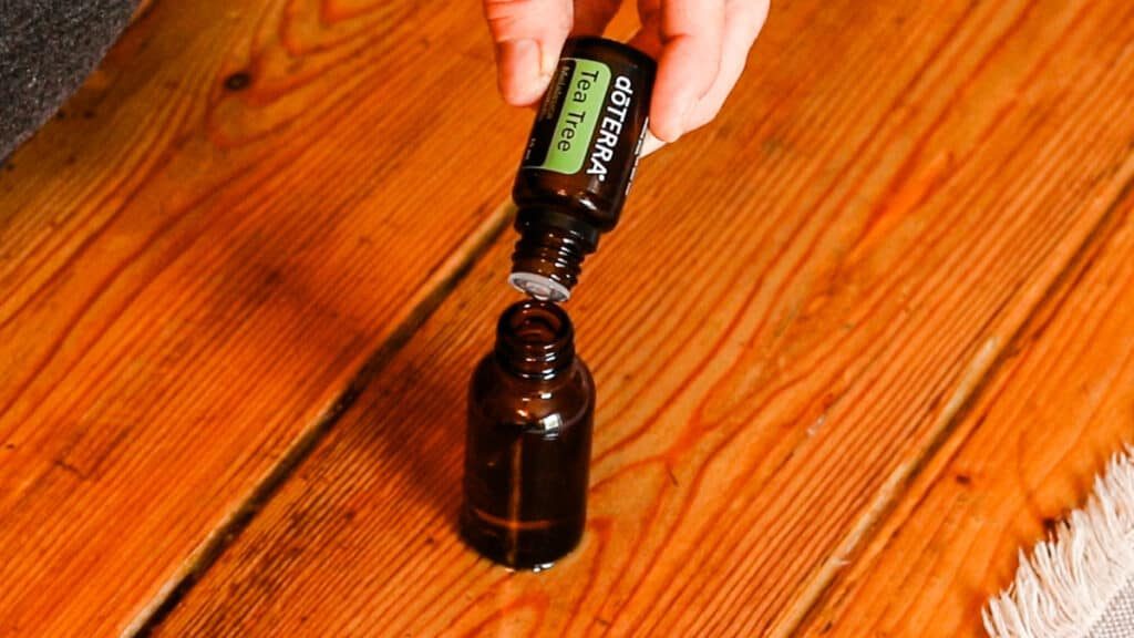 A hand is dropping doTERRA tea tree oil into a brown glass dropper bottle. The bottle sits on a wooden table with an ivory table runner.