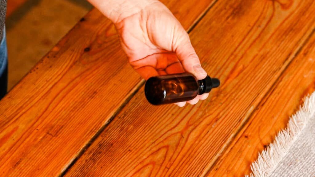 A hand shakes a brown, glass dropper bottle filled with the ingredients for a dry scalp serum. The bottle is being shaken over a wooden table with an ivory table runner.