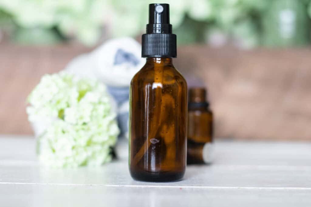 An amber spray bottle of spray deodorant with green flowers, essential oils, and towels in the background.
