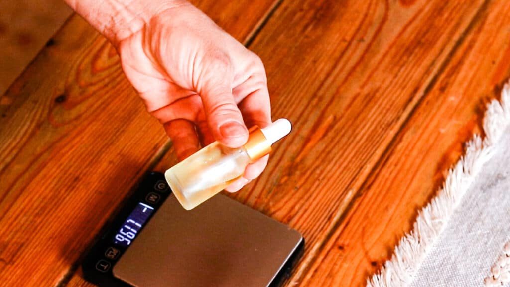 A hand shaking a clear glass bottle with a white dropper to mix up a DIY face serum for youthful skin. There is a digital scale below the bottle over a wooden table.