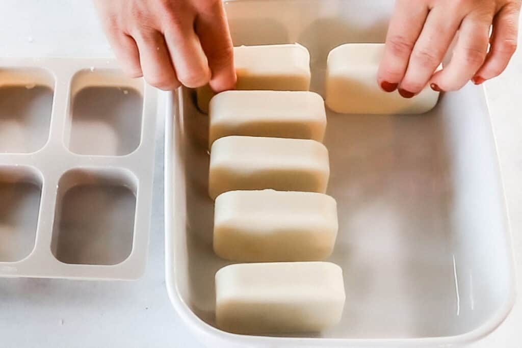 Two hands place white rectangular facial soap bars into a white porcelain tray for curing for 4-6 weeks. The final step in a recipe for cold process facial soap bars.