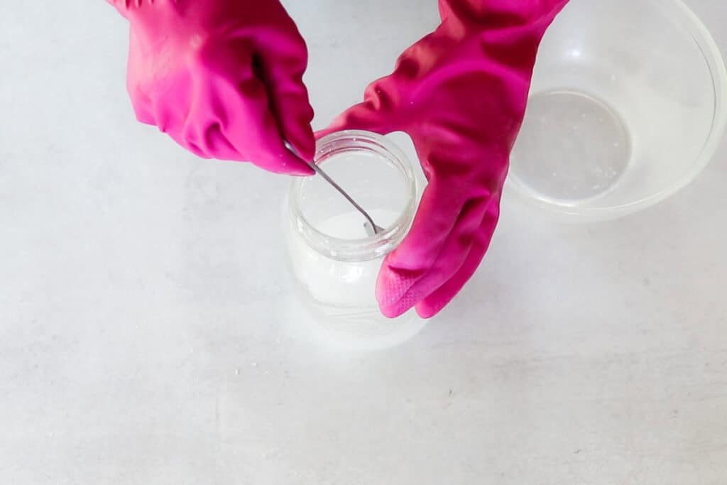 Two hands wearing bright pink rubber gloves for protection are mixing up a lye solution with a silver spoon as a step in making a cold process facial soap recipe.