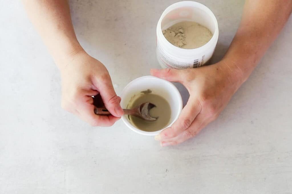 Two hands are holding a bowl and mixing bentonite clay into the water with a silver spoon on a gray countertop. This step is part of a cold process facial soap recipe.