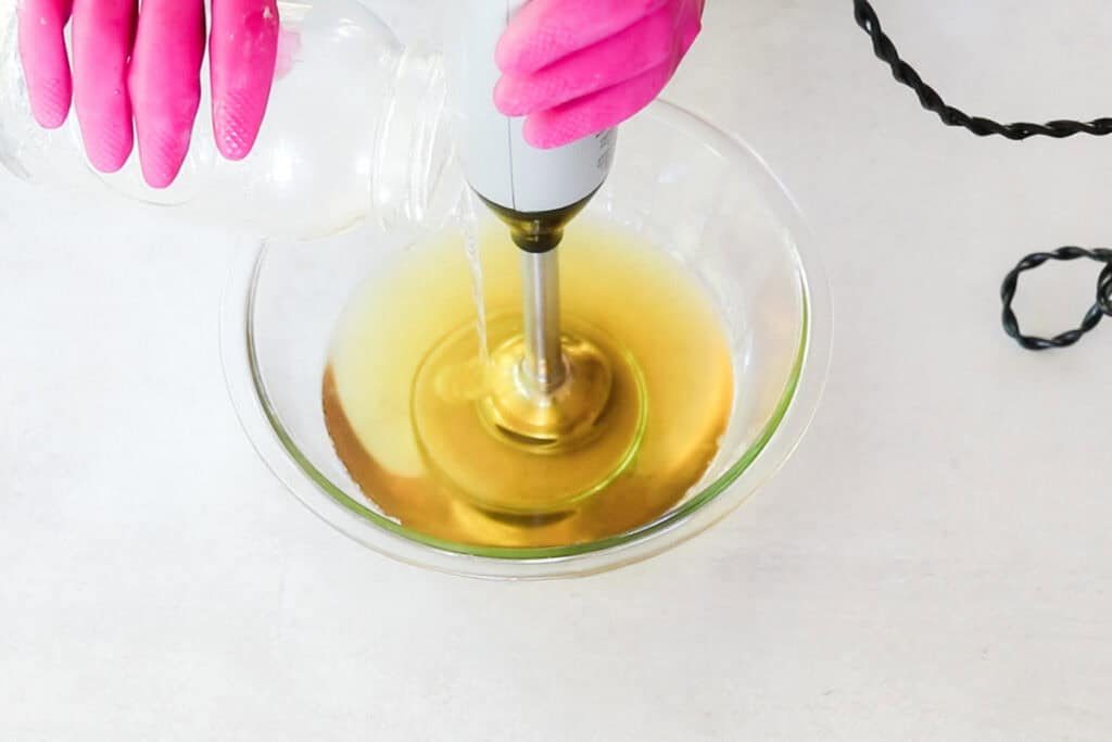 Two hands wearing bright pink rubber gloves use an immersion blender and pour the lye solution into a clear glass bowl filled with melted oils as part of a cold-process facial soap recipe.