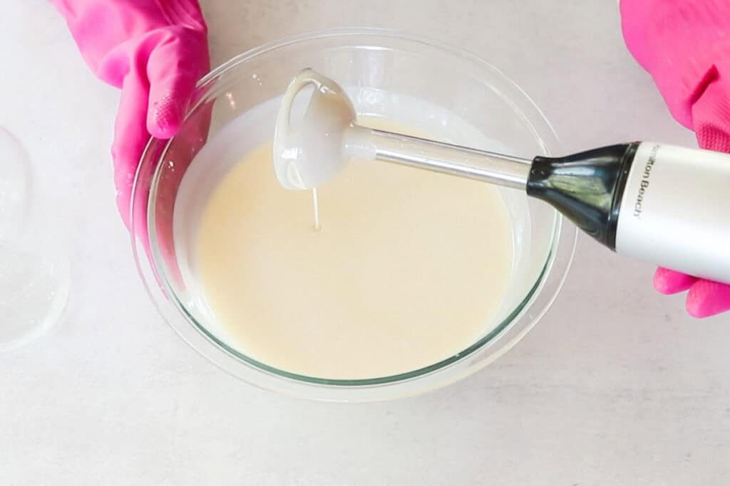 Two hands wearing bright pink rubber gloves use an immersion blender to blend the lye solution with melted oils as part of a cold-process facial soap recipe.