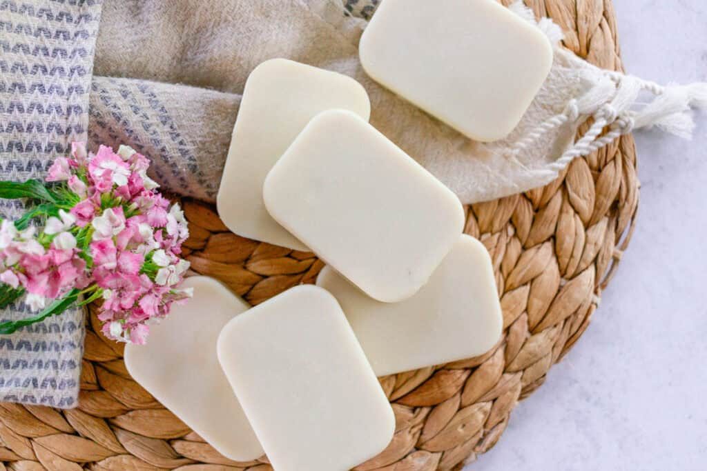 Several white rectangular bars of cold process facial soap lying on a woven placemat. There is a white and blue patterned towel and bunch of pink and white flowers for decoration.