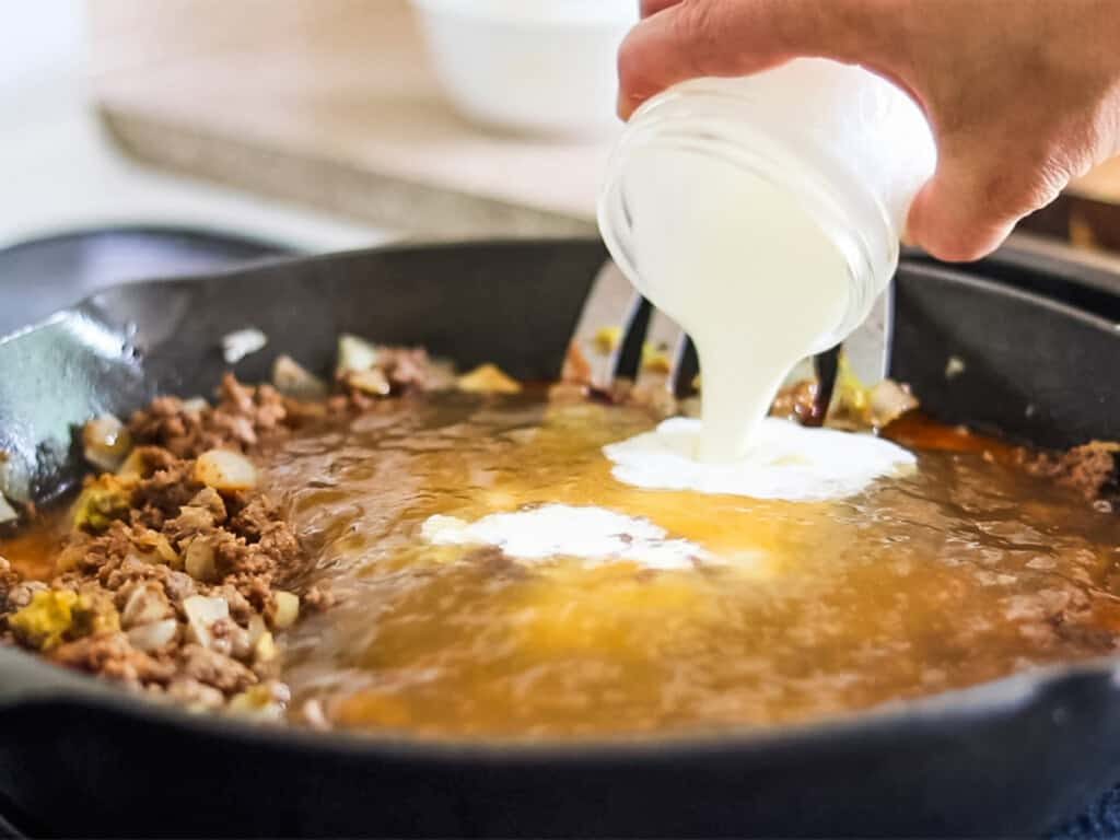 Pouring cream into a skillet with beef and broth to make homemade gluten-free hamburger helper.