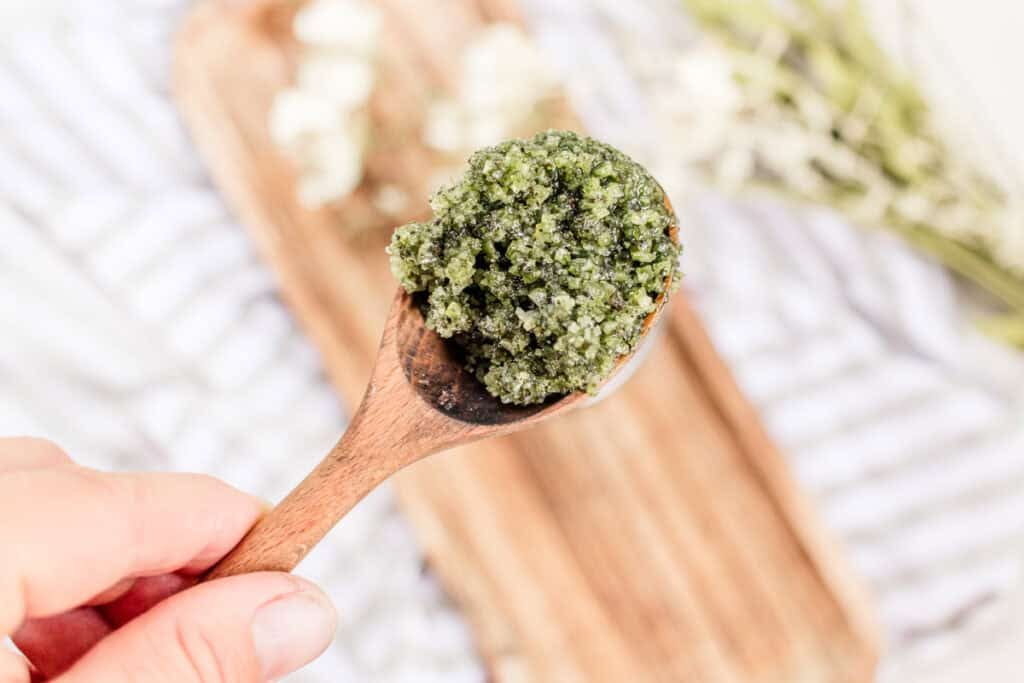 A spoonful of matcha green tea scrub being help up over a wooden cutting board.