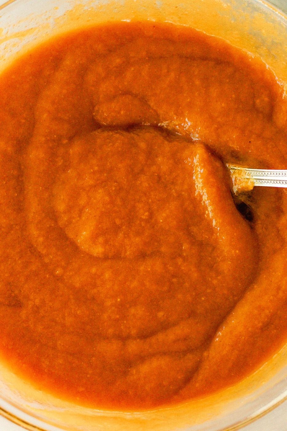Wet ingredients for the pumpkin muffins getting stirred in a clear mixing bowl with a metal spoon.