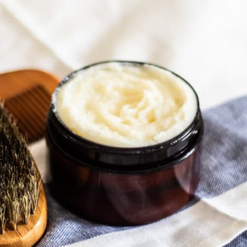A jar of beard conditioning balm on a striped towel with a brush.