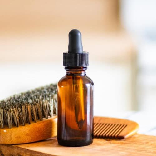 A bottle of homemade beard oil with a beard brush on a wooden board.