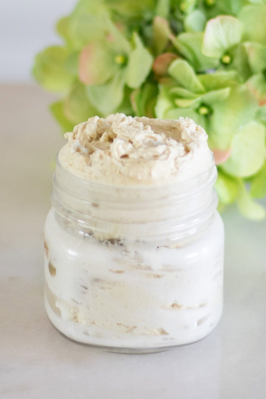 A glass jar of homemade body butter with green flowers in the background.