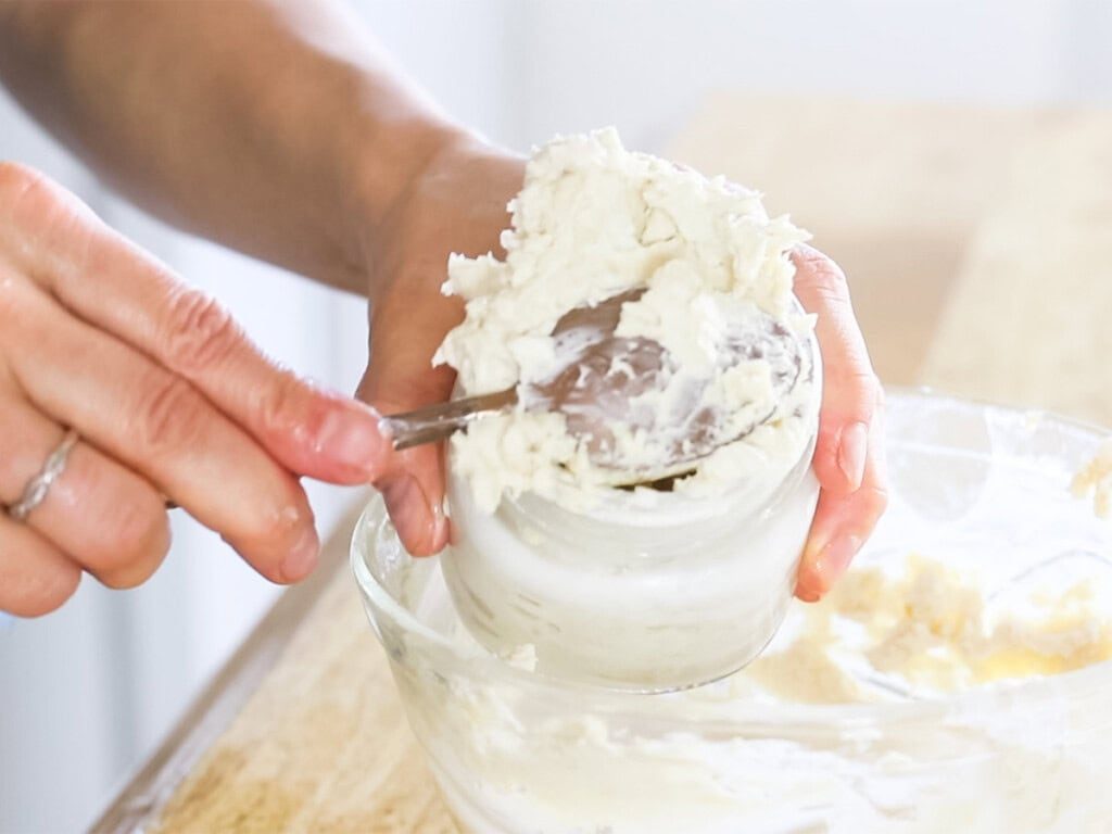 Scooping homemade body butter into a glass jar with a spoon.