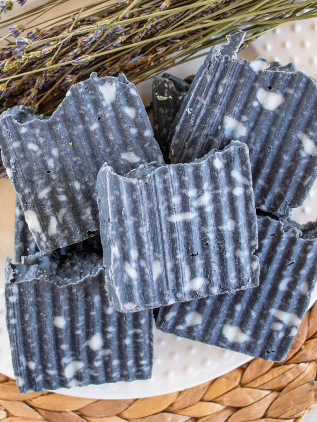 Bars of homemade charcoal soap stacked up on a white plate and a wicker placemat.