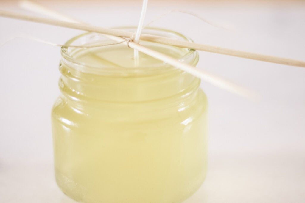 A jar of melted wax with a wick sticking out of it, wrapped around a stick to hold it up.