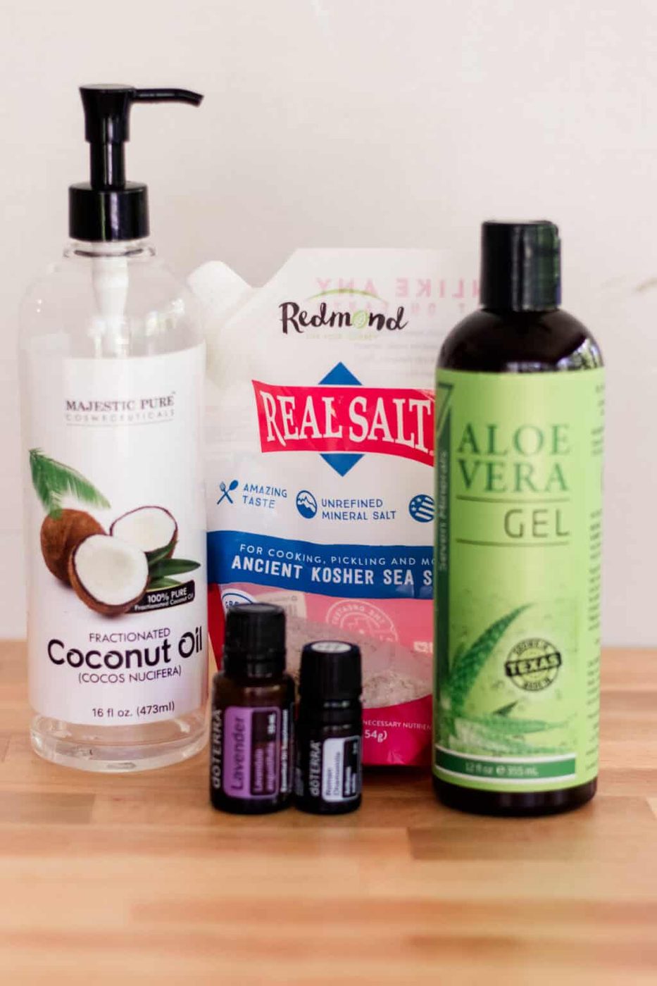 A bottle of coconut oil, a bottle of aloe vera, a bag of sea salt, and two bottles of essential oils assembled on a countertop.