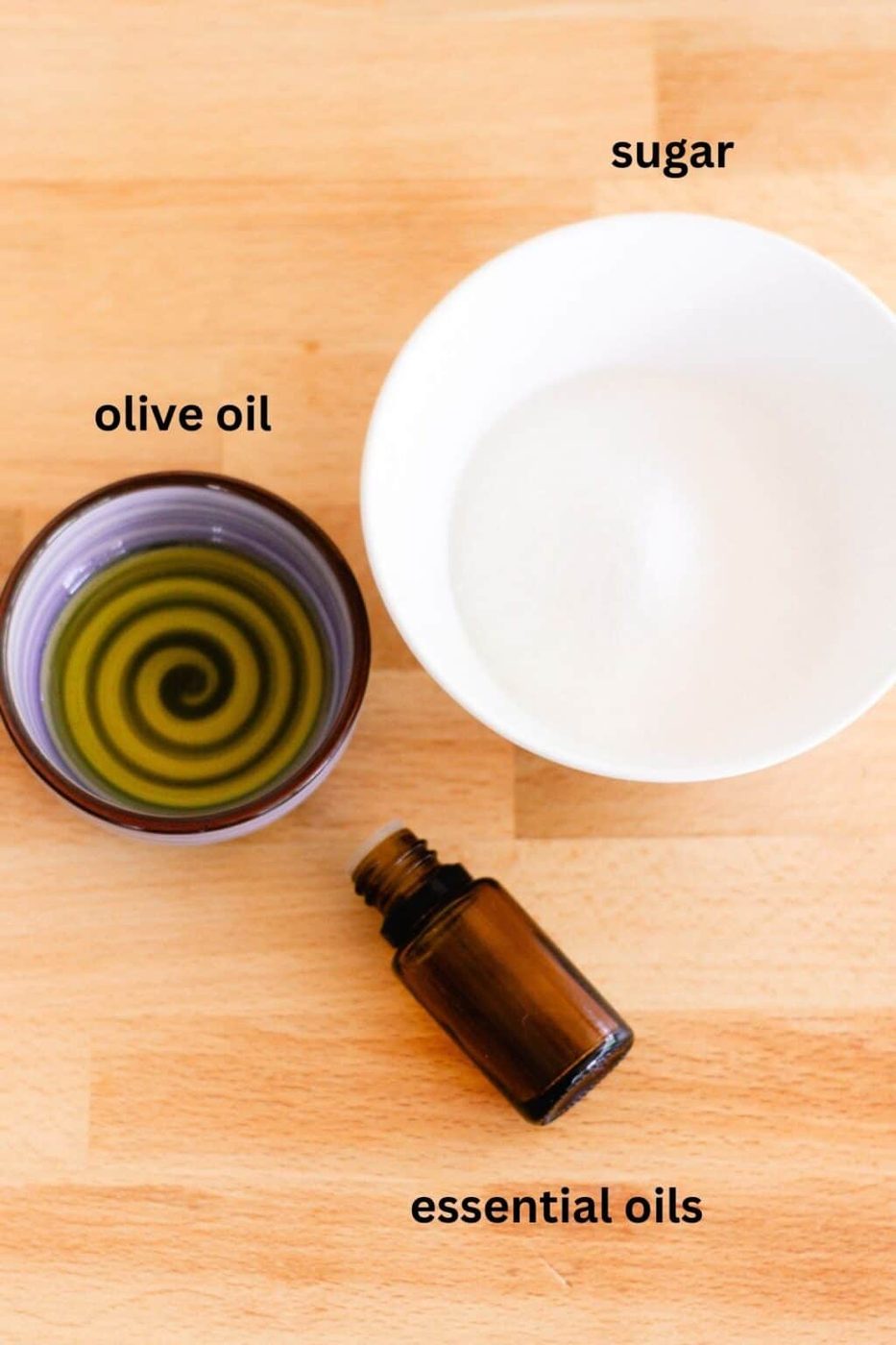 Ingredients laid out for a homemade sugar scrub. Olive oil, sugar, and a small bottle of essential oil.