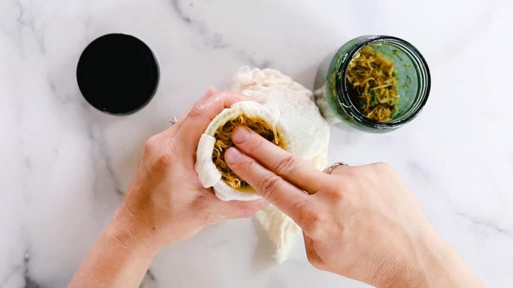 A person is using two fingers to smash the calendula petals into the cheesecloth to strain homemade calendula oil into a small jar. The black lid rests on the white countertop.