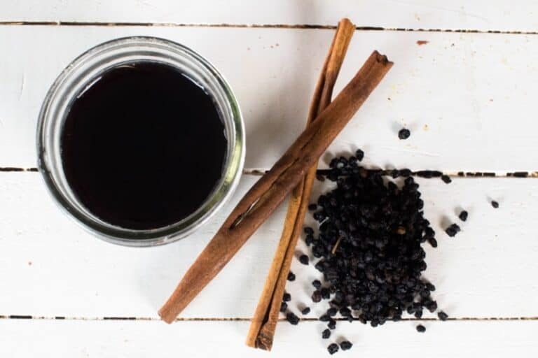 A jar of elderberry syrup on a white table with cinnamon sticks and dried elderberries.