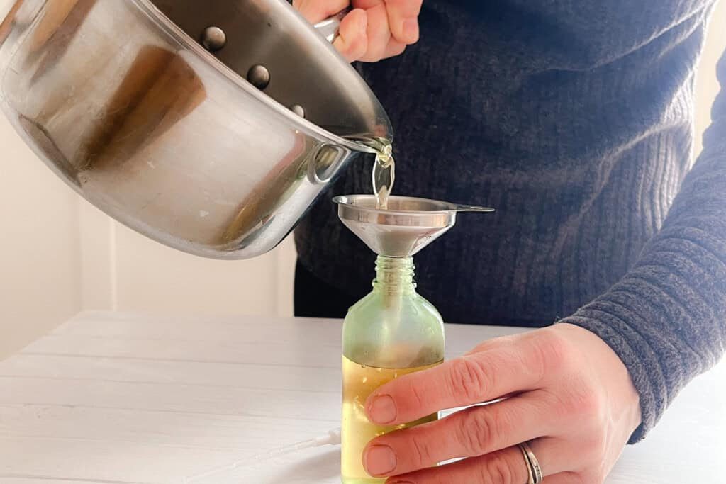Pouring the cooled rosemary water into a clear spray bottle using a metal funnel.