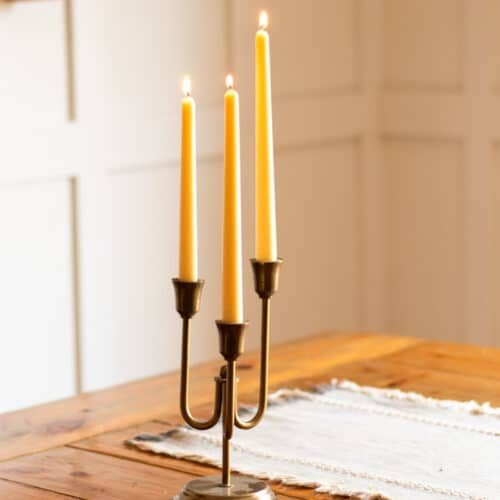 Three beeswax taper candles burning in a bronze candle holder on a wooden table.