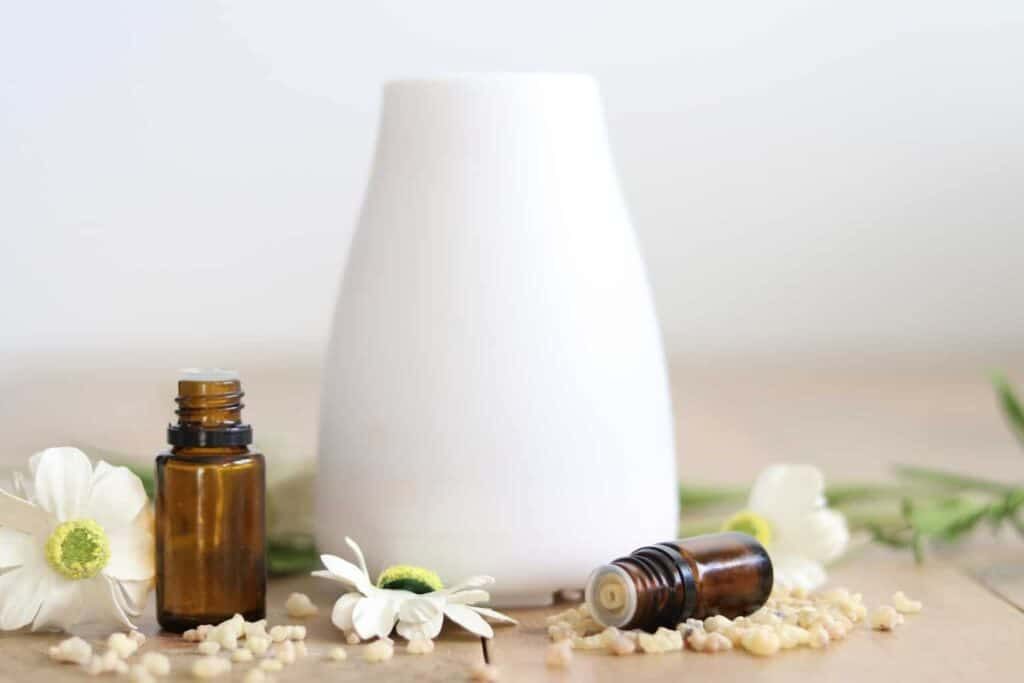White essential oil diffuser surrounded by flowers and oil bottles on wooden table.
