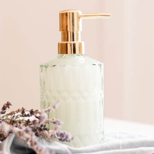 Homemade liquid hand soap in a green tinted embossed glass soap dispenser on top of a folded towel with dried flowers lying beside it.