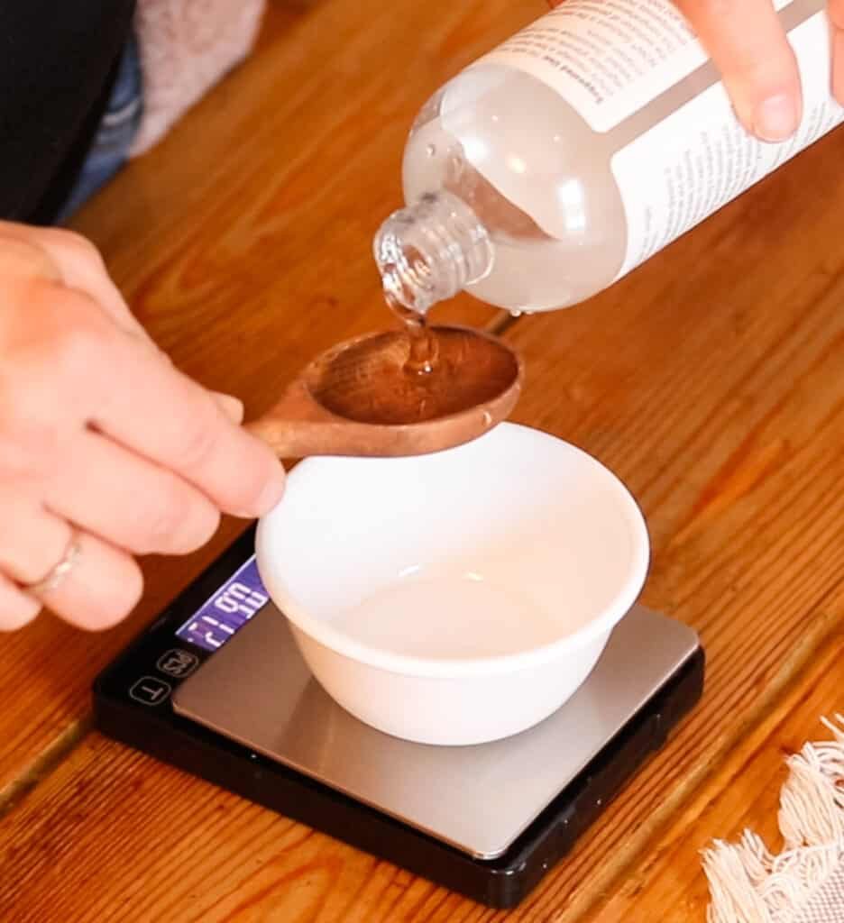 Pouring vegetable glycerin into a wooden spoon held over a small bowl that is sat on top of a digital scale.