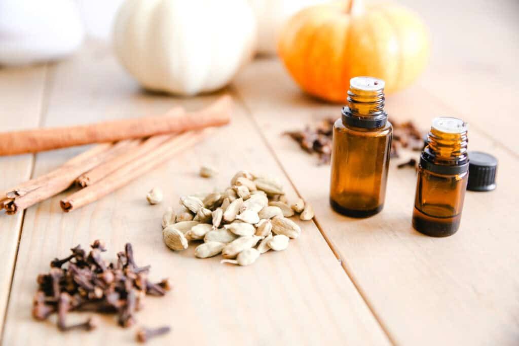 An assortment of spices and essential oil bottles on a wooden table with pumpkins in the background.