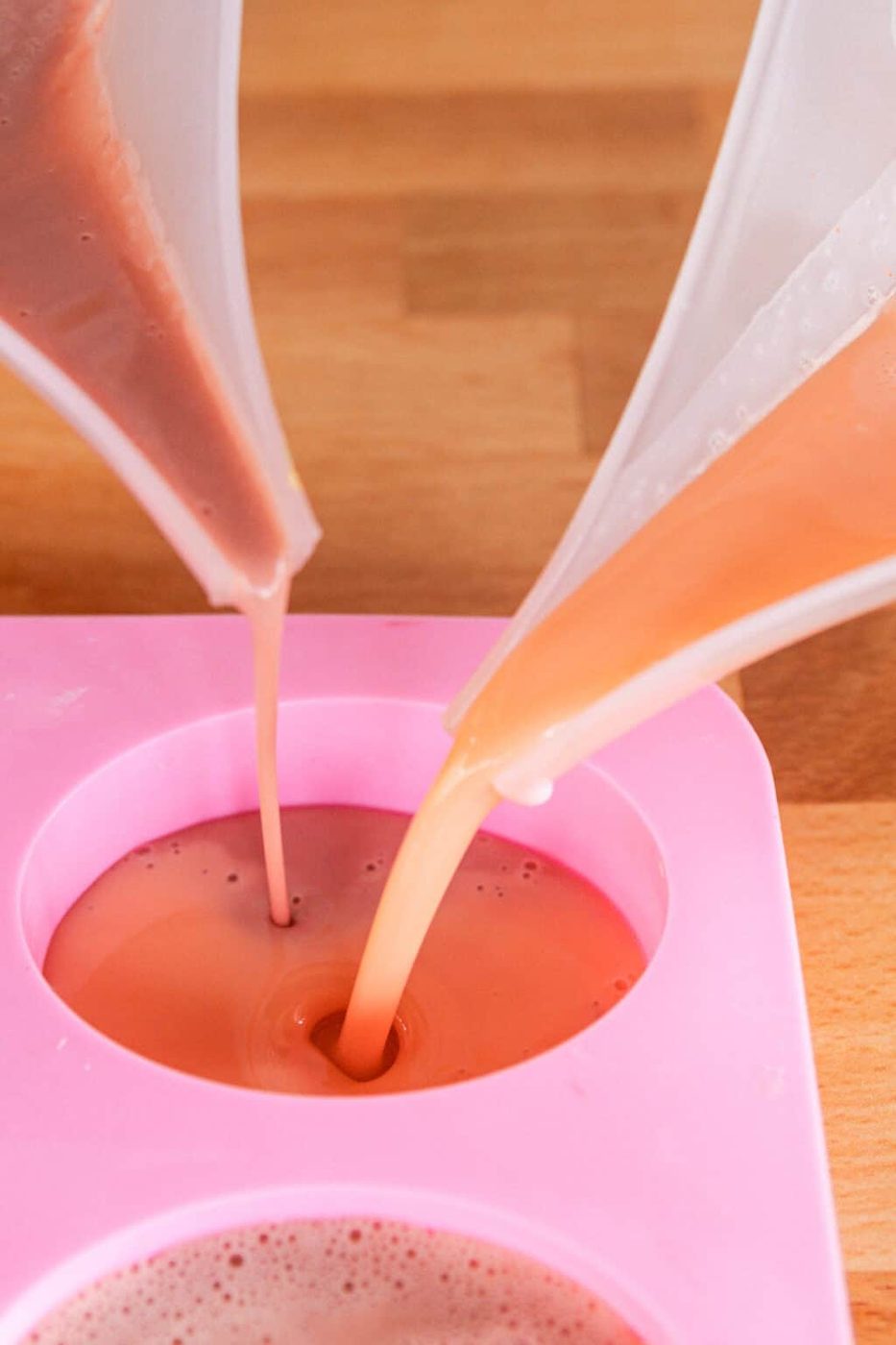 Pouring two different colors of soap base into a circular silicone mold.
