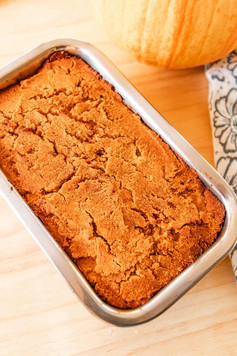 Paleo pumpkin bread cooling in a loaf pan on a wooden table with a decorative flower towel next to it.