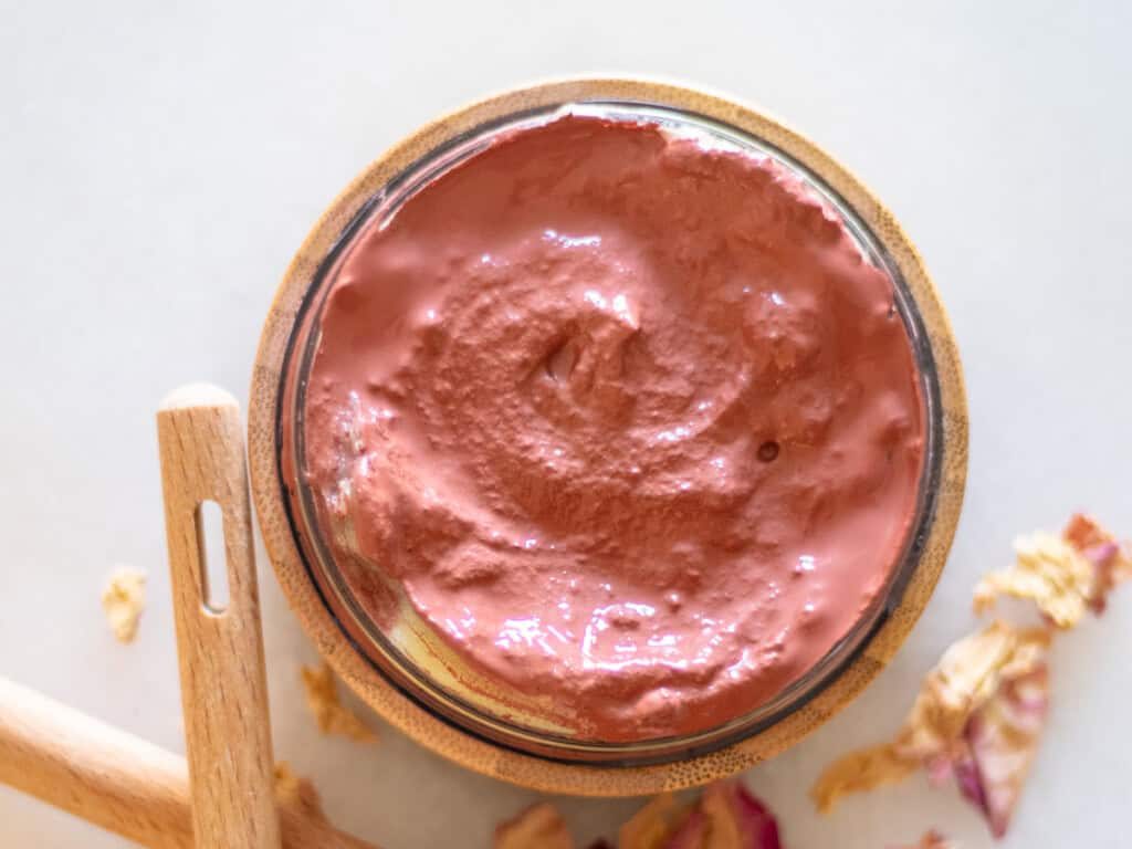 A jar of homemade rose clay face mask with rose petals around it.