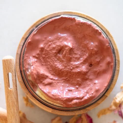 A jar of homemade rose clay face mask with rose petals around it.