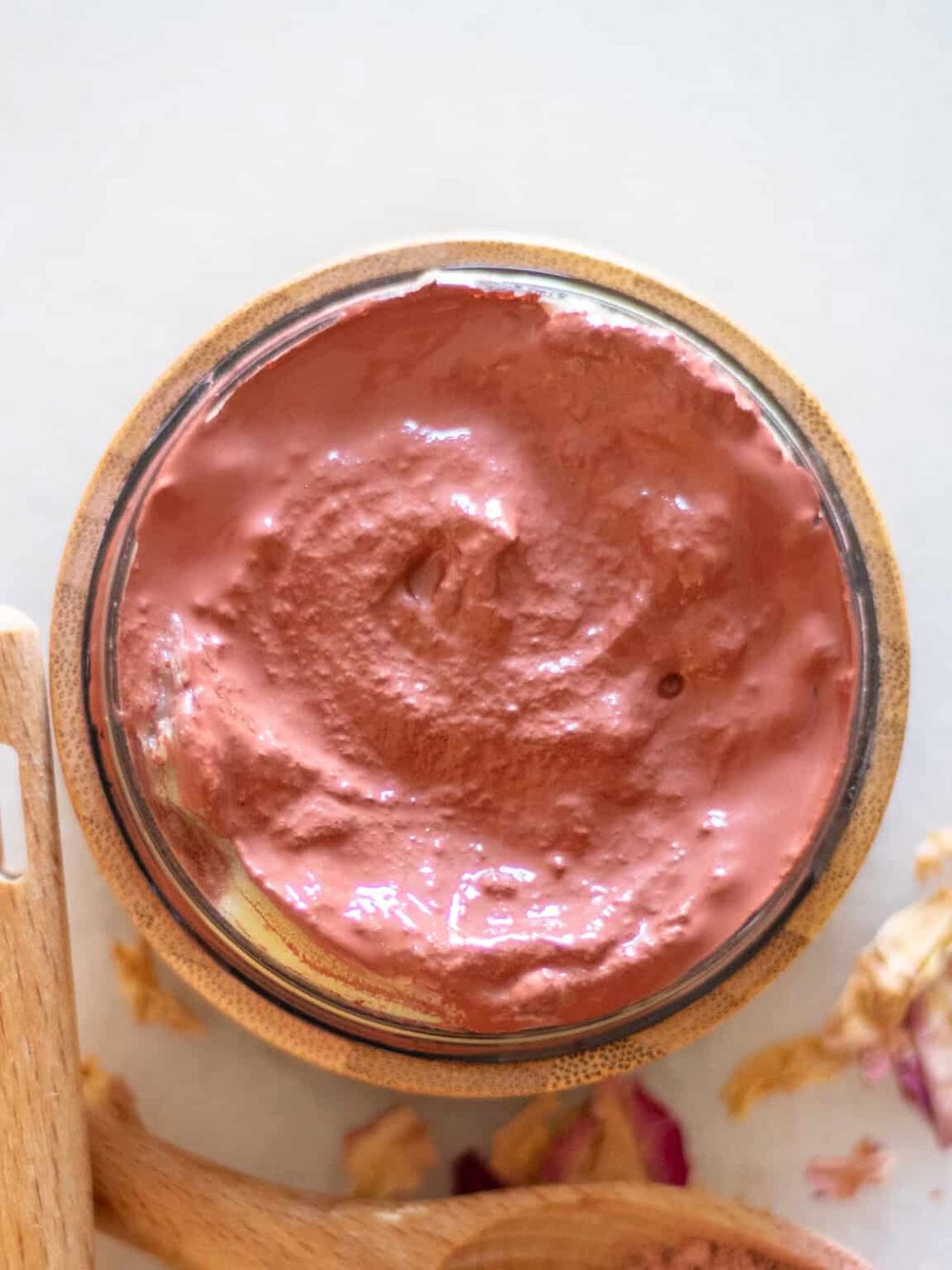A bowl of rose clay face mask with rose petals and a wooden spoon.