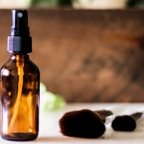DIY makeup setting spray in glass spray bottle with applicator brushes on white marble vanity.