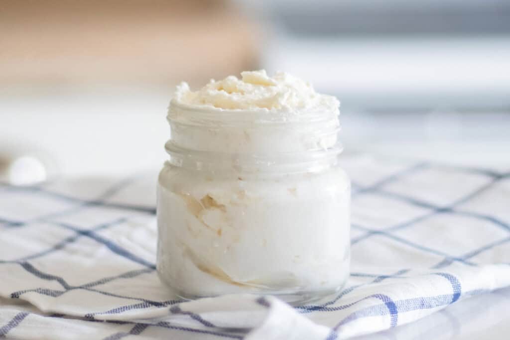 A jar of homemade shea butter lotion on a white and blue checkered tablecloth.
