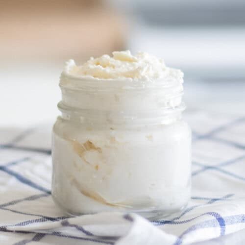 A jar of homemade shea butter lotion on a white and blue checkered tablecloth.